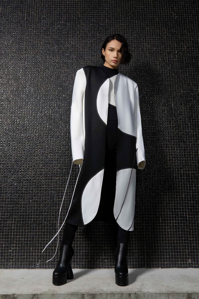 The female model wears the oversized Alexi neoprene coat with contrasting organic cutouts on the front and back. Ultra-long sleeves with lace up. The outfit is paired with a pair of black ankle boots and leggings.